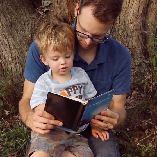 A man and a young child are sitting outdoors, reading a storybook together under a tree.
